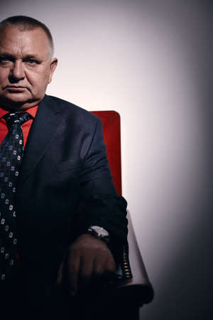 Portrait of serious middle aged businessman wearing black suit, red shirt and wristwatch sitting on old fashioned sofa in officeの写真素材