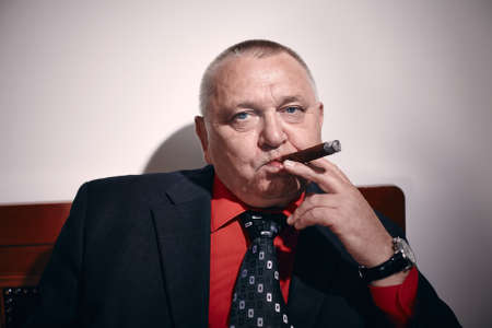 Portrait of serious middle aged businessman wearing black suit, red shirt and wristwatch sitting on old fashioned sofa in office and smoking cigarの写真素材