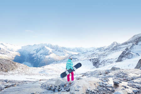 Back view of female snowboarder wearing colorful helmet, blue jacket, grey gloves and pink pants standing with snowboard in one hand and enjoying alpine mountain landscape - winter sports conceptの写真素材