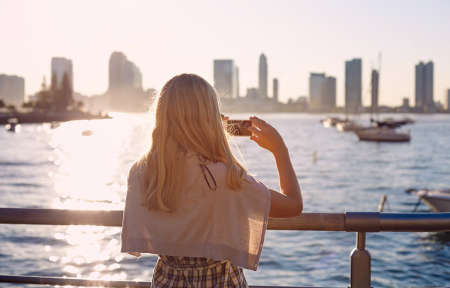 Blonde girl in glasses taking picture of city bay with mobile phone on sunset. Focus on smartphoneの写真素材