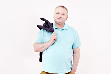 Mature cheerful man wearing blue shirt and brown trousers standing with hand in pocket, holding blue...