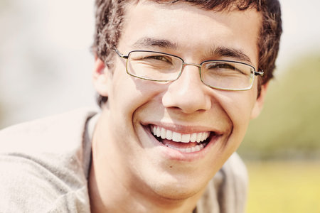 Warm toned close up portrait of young hispanic man wearing glasses, smiling perfect healthy toothy smile in spring park outdoors - humor, dentistry or ophthalmology conceptの写真素材