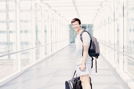 Young hispanic man wearing glasses, t-shirt, shorts and backpack, standing in airport corridor, holding mobile phone and sport handbag in his hands, looking at camera and smiling - travel conceptの写真素材