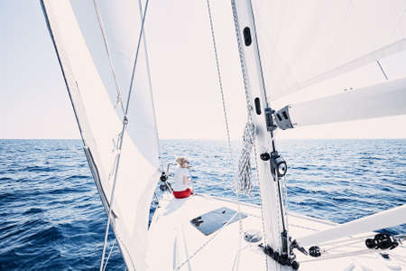 Young blonde woman wearing sunglasses and red shorts, sitting on deck under sails on yacht bow and enjoying wonderful view to peaceful sea during summer sailing holidays - yacht charter conceptの写真素材