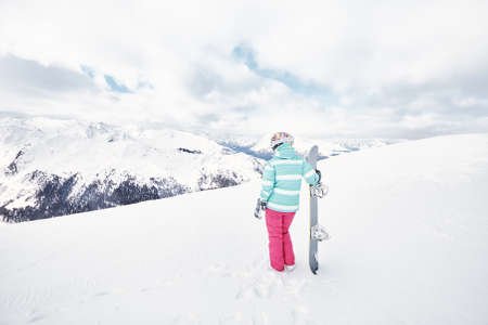 Back view of female snowboarder wearing colorful helmet, blue jacket, grey gloves and pink pants standing with snowboard in one hand and enjoying alpine mountain landscape - snowboarding conceptの写真素材