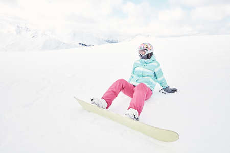 Female snowboarder wearing colorful helmet, blue jacket, grey gloves and pink pants sitting with yellow snowboard on snow and preparing for ride - snowboarding conceptの写真素材