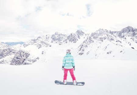 Female snowboarder wearing colorful helmet, blue jacket, grey gloves and pink pants standing against snowy mountains and preparing for ride - snowboarding conceptの写真素材
