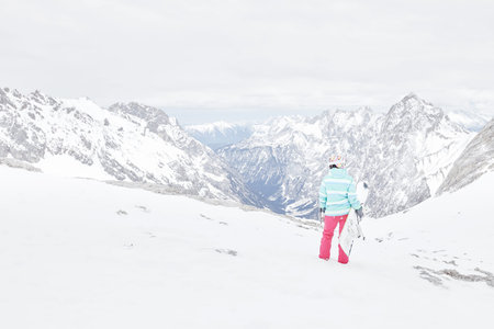 Back view of female snowboarder wearing colorful helmet, blue jacket, grey gloves and pink pants standing with snowboard in one hand and enjoying alpine mountain landscape - snowboarding conceptの写真素材