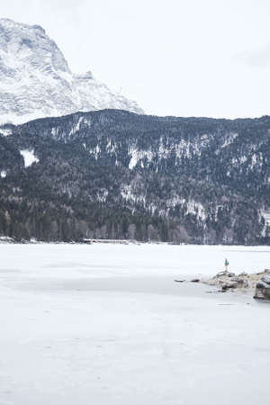 Young female hiker with backpack enjoying view to frozen lake Eibsee, winter Bavarian forest and snowy mountains at base of Zugspitze, Alps - nature lovers travel conceptの写真素材