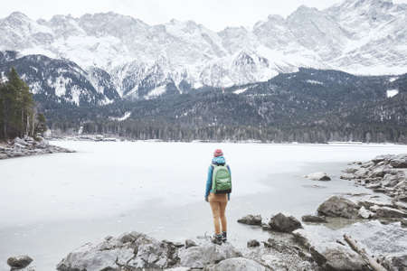 Young female hiker with backpack enjoying view to frozen lake Eibsee, winter Bavarian forest and snowy mountains at base of Zugspitze, Alps - nature lovers travel conceptの写真素材