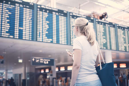 Young blonde woman with phone in her hand and shoulder bag checking flight timetable in international airport - travel conceptの写真素材