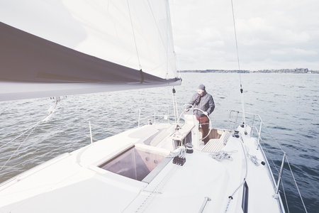 Senior man wearing jacket, scarf and hat, standing on yacht steering wheel and enjoying perfect...