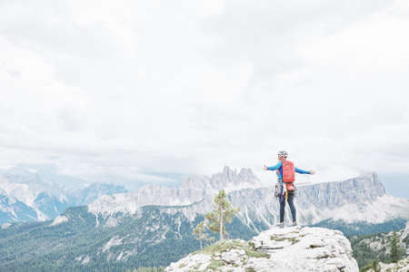 Female mountaineer with backpack, helmet and harness with climbing gear standing on top of mountain with arms outstretched and enjoying view to valley and peaks in Dolomite Alps - freedom conceptの写真素材