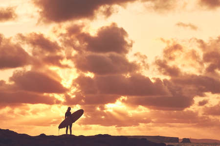 Young woman wearing shorty wetsuit, standing on beach with surfboard in her hands and enjoying stunning sunset after evening surfing session - water sports concept. Baleal, Peniche, Portugalの写真素材