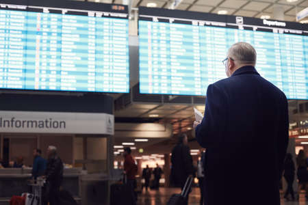 Back view of adult man wearing black coat, holding tickets and passport in hand, checking flight timetable in international airport - business travel without delays conceptの写真素材