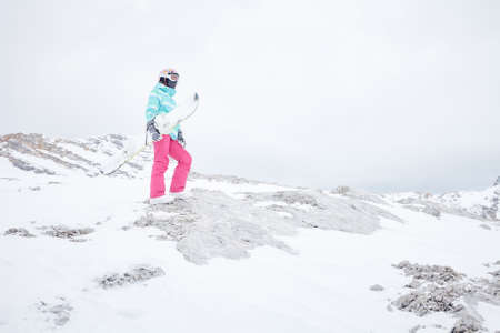 Female snowboarder wearing colorful helmet, blue jacket, grey gloves and pink pants standing with snowboard in one hand and enjoying alpine mountain landscape - snowboarding concept, copy spaceの写真素材