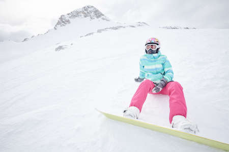 Female snowboarder wearing colorful helmet, blue jacket, grey gloves and pink pants sitting with snowboard on snow and preparing for ride - snowboarding conceptの写真素材