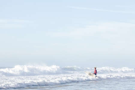 Young adult surfer with dreadlocks wearing wetsuit entering foggy ocean with surfboard under arm to...