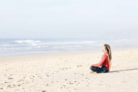 Young adult surfer with dreadlocks wearing wetsuit sitting cross-legged on beach near spot and meditating before summer evening surfing session - yoga and surfing concept. Baleal, Peniche, Portugalの写真素材