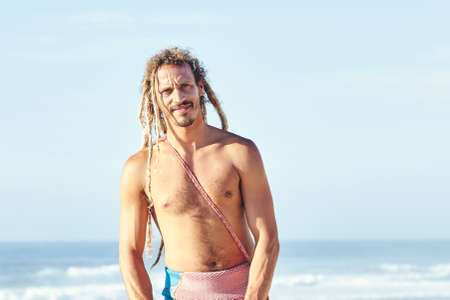 Portrait of young adult shirtless surfer with dreadlocks standing and smiling on beach against surfing spot before summer evening session - surfing and extreme sport concept. Baleal, Peniche, Portugalの写真素材
