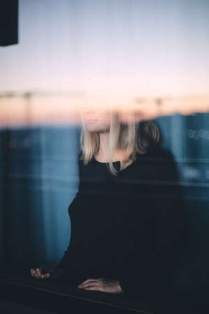 Mood portrait of young blonde woman wearing glasses looking through window enjoying cityscape at sunset - calm and relax in city conceptの写真素材