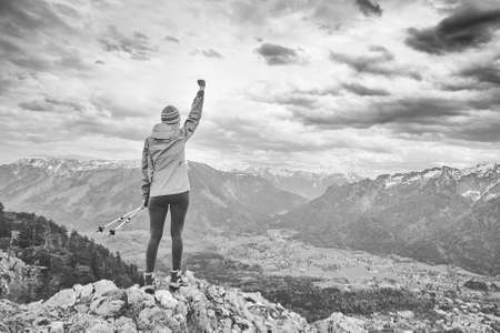Black and white portrait of young female hiker with trekking poles and arm raised celebrating achievement standing on top of mountain after successful ascent - adventure, freedom or success conceptの写真素材