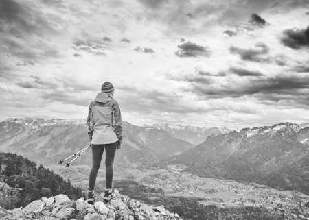 Black and white portrait of young female hiker with trekking poles standing on top of mountain and enjoying stunning view to valley under dramatic sky - adventure, hiking and active lifestyle conceptの写真素材