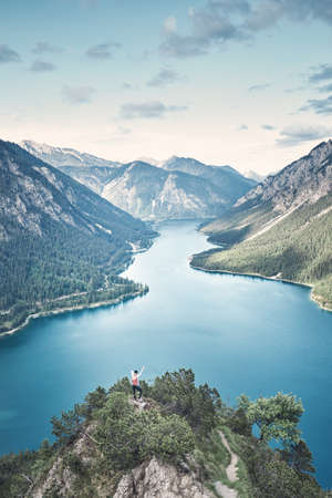 Happy female adventurer with backpack and arm raised enjoying amazing view to mountain lake in valley from top of mountain after summer hike - adventure, travel and hiking concept. Plansee, Austriaの写真素材