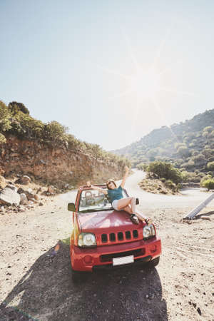 Happy beautiful carefree young woman having fun sitting on bonnet of convertible 4x4 car in mountains - road trip adventure conceptの写真素材
