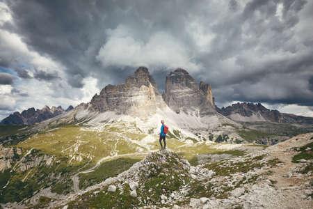 Female hiker with red backpack enjoying stunning view to Tre Cime di Lavaredo (Drei Zinnen) under heavy clouds from hiking trail during hike in Dolomites, Italy - adventure, travel and trekkingの写真素材