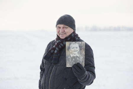 Senior man wearing knit cap, scarf, jacket and leather gloves smiling and showing his picture as child standing in winter field - memory conceptの写真素材