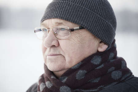 Close up portrait of senior man wearing glasses, knit cap and scarf standing outdoors in winterの写真素材