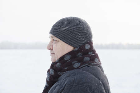 Profile portrait of senior man wearing knit cap, scarf and jacket standing in winter field during snowfallの写真素材