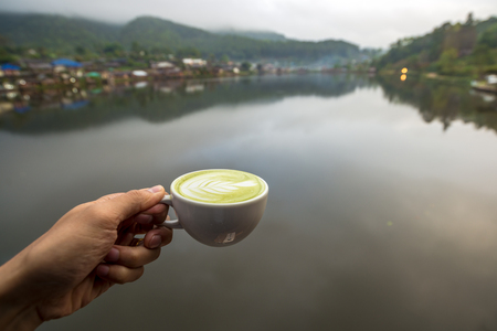 Hot Matcha latte green tea in white cup on hand with water village at the edge of river view, Traveling in Thailandの写真素材