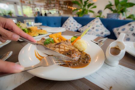 Cutting the steak by knife in white dish while take a dinner, to enjoy with the main course