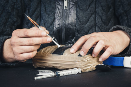 Male hand holding in the palm of a plastic bag with powder cocaine or other drugs and a syringe. On...