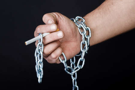 Cigarettes and hand of a man tangled in chains . The concept of nicotine addiction. Isolated on black background.の写真素材