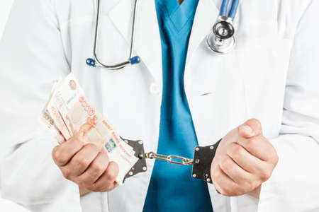 Doctor with a stethoscope and 5000 banknotes and handcuffs. a crime. on a white background.の写真素材