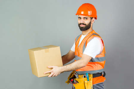 A man-builder in an orange helmet with a cardboard box in his hands. On a gray background.の写真素材