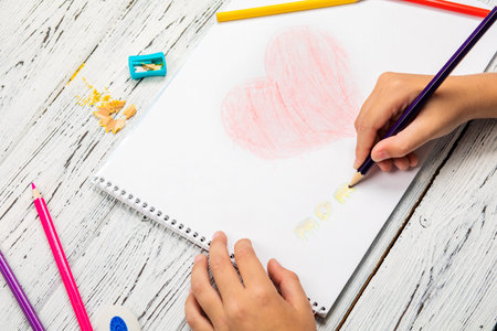 Hand of children drawing red heart with colored pencil on white paper on wooden table