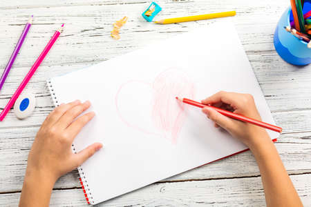 Hand of children drawing red heart with colored pencil on white paper on wooden tableの写真素材