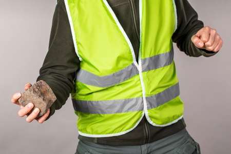 Rebel or protesting worker in a yellow vest with a brick in his hands on a gray background.の写真素材