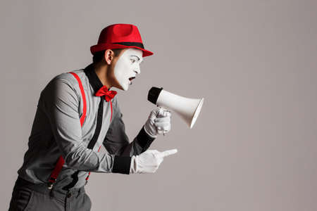 The clown, MIM, is holding a Megaphone. Expression of emotions. On a gray background.