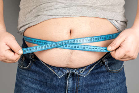 A man measures his fat belly with a measuring tape. on a gray backgroundの写真素材