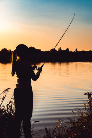 Silhouette of a woman at sunset with a fishing rod near the pondの写真素材