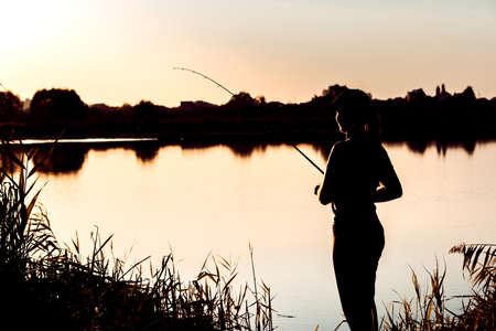 Silhouette of a woman at sunset with a fishing rod near the pondの写真素材