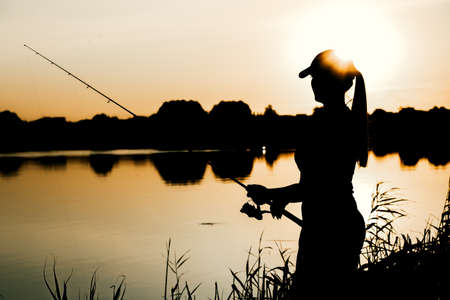 Silhouette of a woman at sunset with a fishing rod near the pond