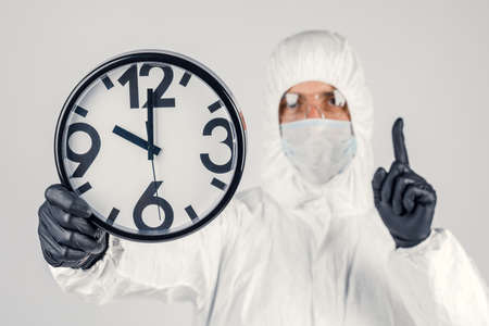 A man in a white protective suit , a mask on a white background, with a watch in his hands. An...