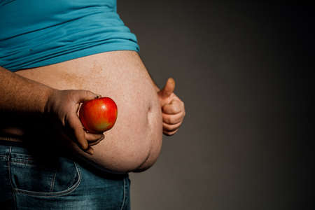 The fat man holds a Apple in front of his bare stomach. The concept of diet and healthy eating.の写真素材