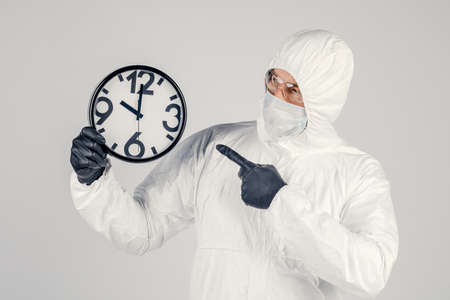 A man in a white protective suit , a mask on a white background, with a watch in his hands. An epidemic pandemic is a new rapidly spreading virus, a medical conceptの写真素材